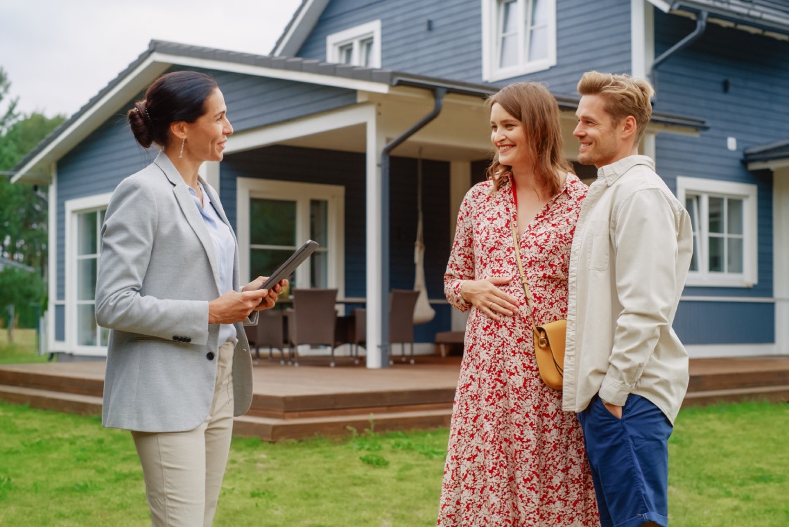 Young Couple Visiting a Potential New Home Property with Professional Real Estate Agent. Female Realtor Showing the House to Future Homeowners. Focus on For Sale Sign.