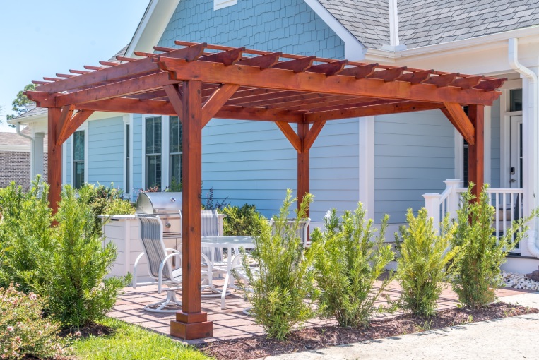 Trendy outdoor patio pergola shade structure made of dark wood, awning and patio roof, white dining table, white chairs, metal grill surrounded by landscaping in a luxury American neighborhood