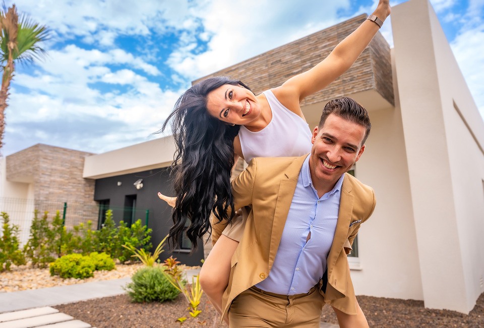 A joyful couple, with the woman on the man's back, poses playfully outside a modern home surrounded by greenery. New home owners. 