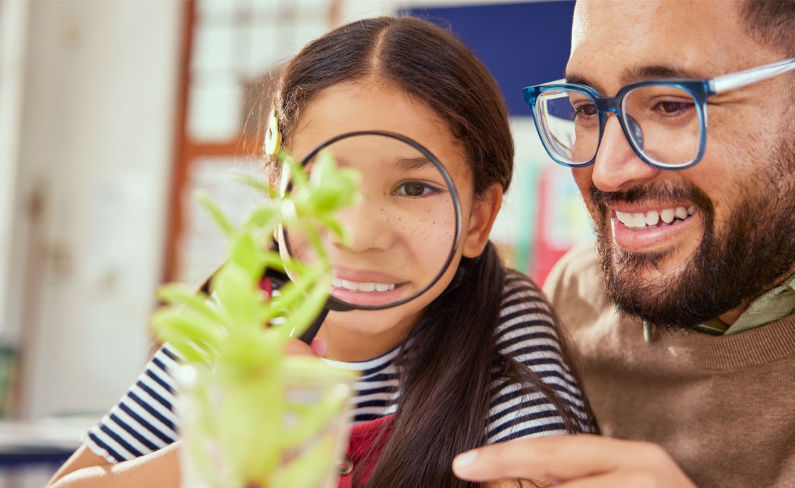 Cheerful male teacher helping child understand plants using magnifying glass in classroom. Curious cute little girl with magnifying glass learning about plants during biology lesson at elementary.