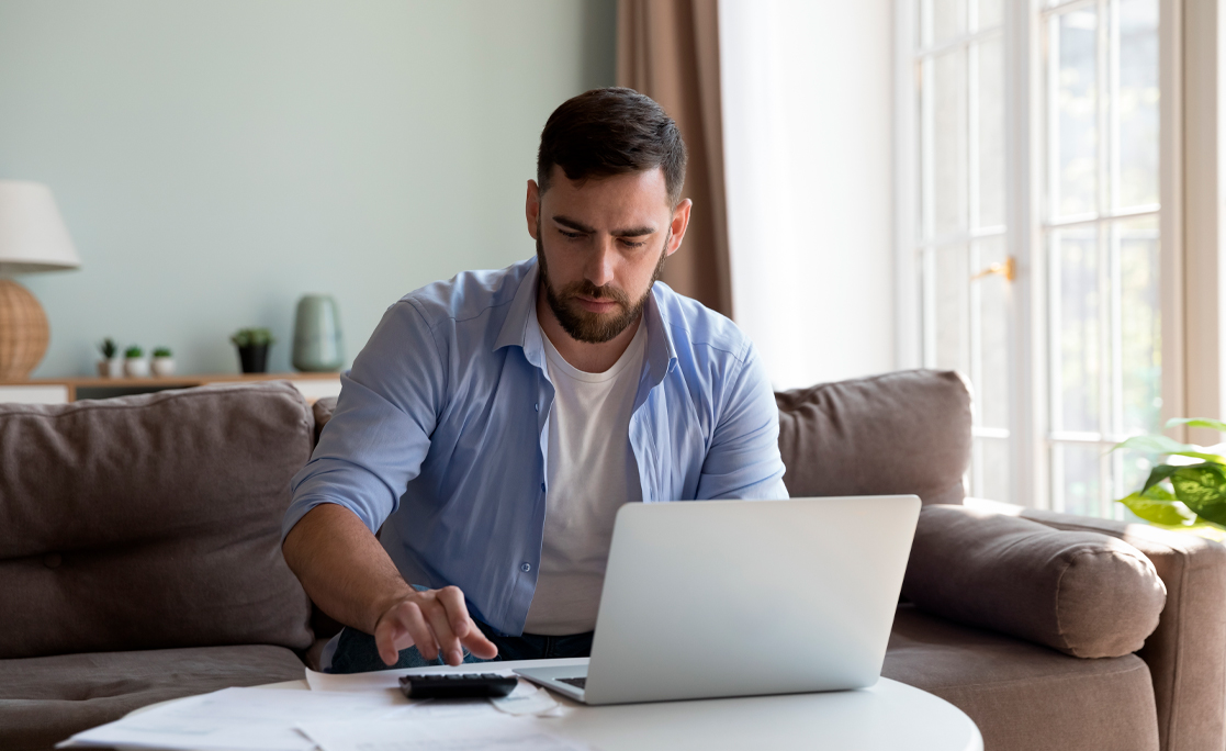 Serious millennial man sit on sofa do paperwork, pay taxes and household utility bills on-line using e-bank app on laptop, manage budget, planning savings, investments looks focused. Finances